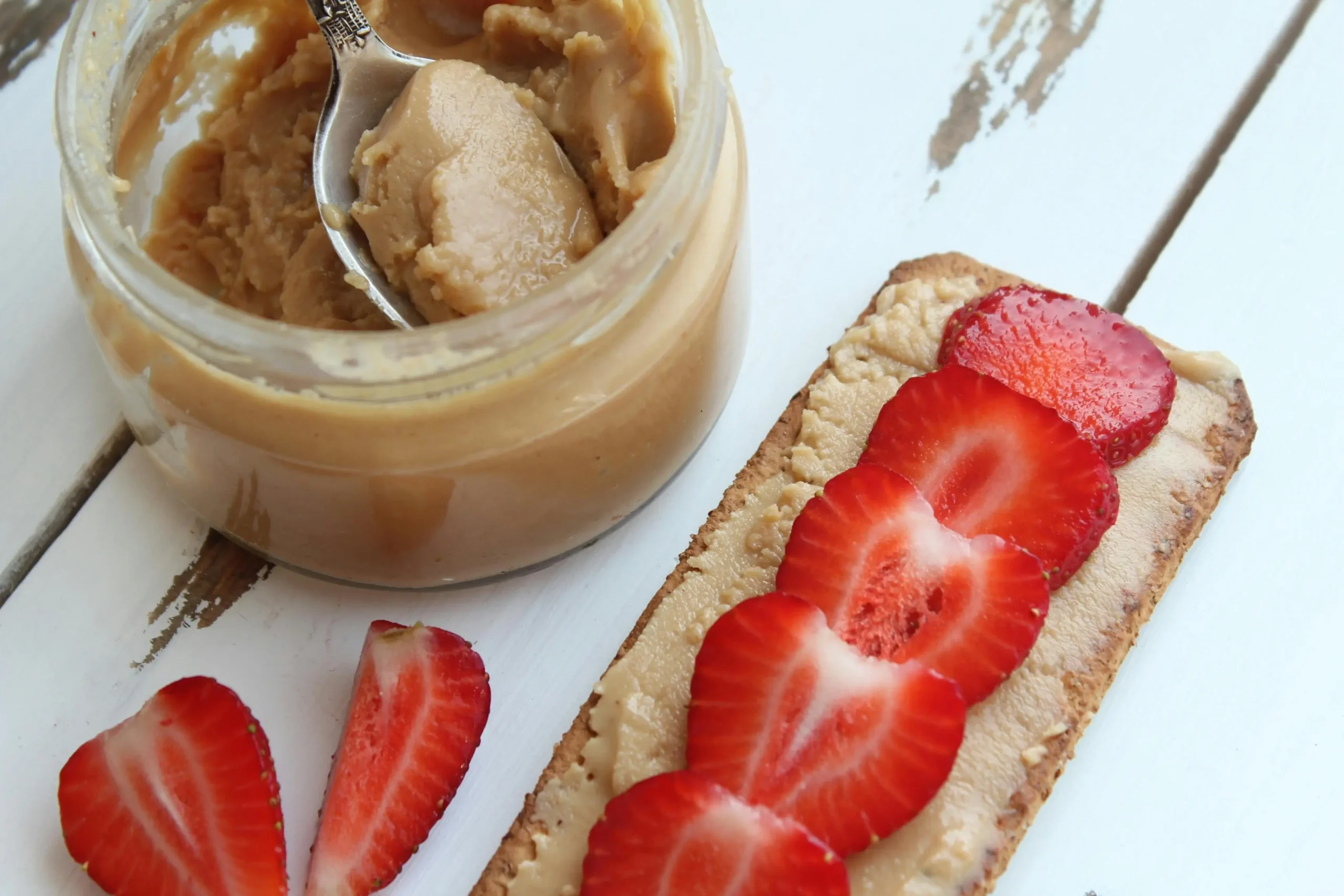 A jar of creamy peanut butter with a spoon dipped in, surrounded by whole peanuts and served with fresh bread slices for a wholesome snack.